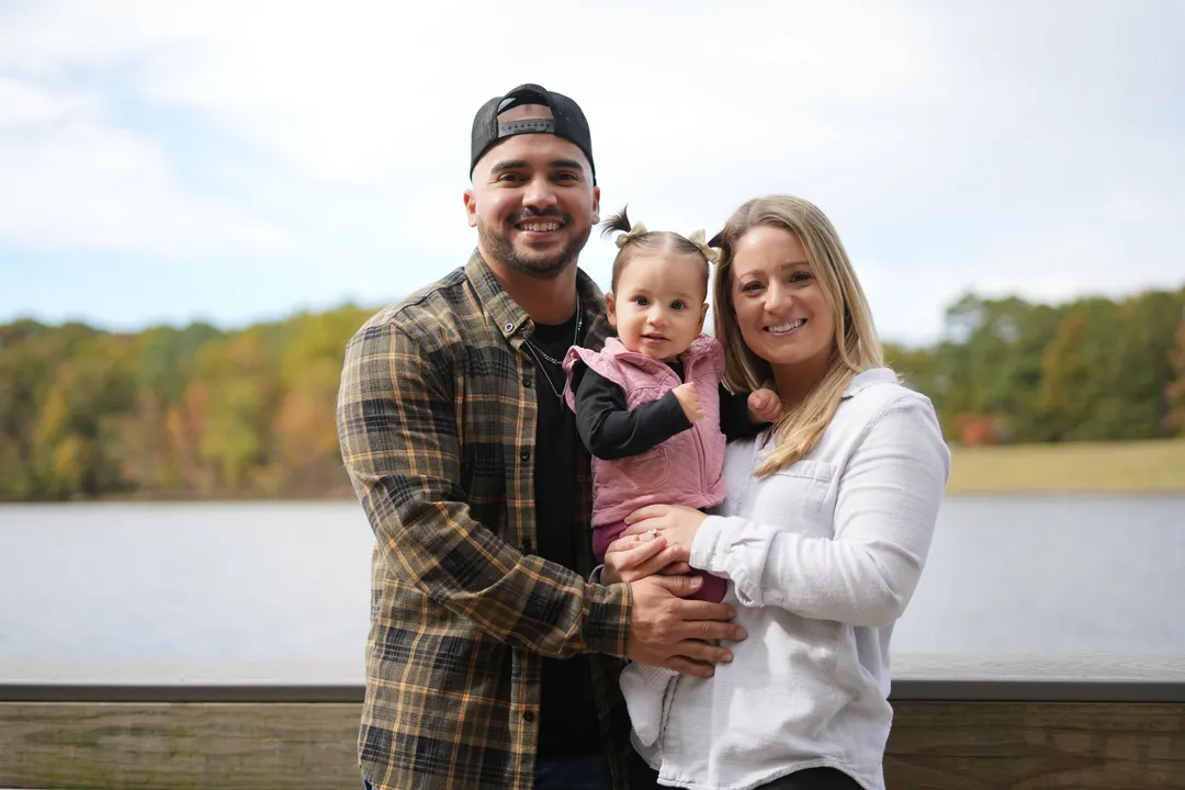 Mario and Ashley smiling by the lake with baby Sloan between them