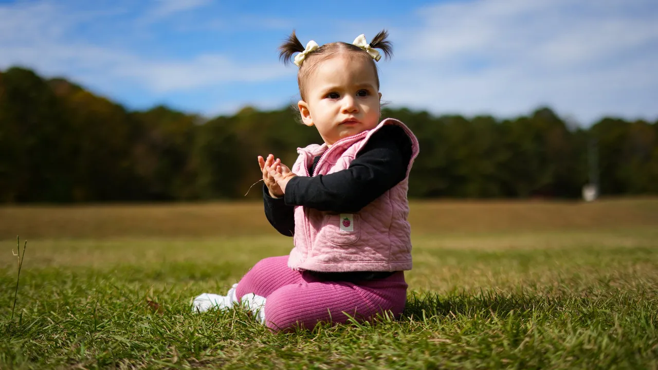 Sloan sitting in the grass, clapping, in a pink vest and pigtails